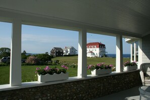 A covered porch with stone walls and white columns, overlooking a grassy area with a large white building and a red-roofed structure in the distance. - Rose Farm Inn (Block Island)