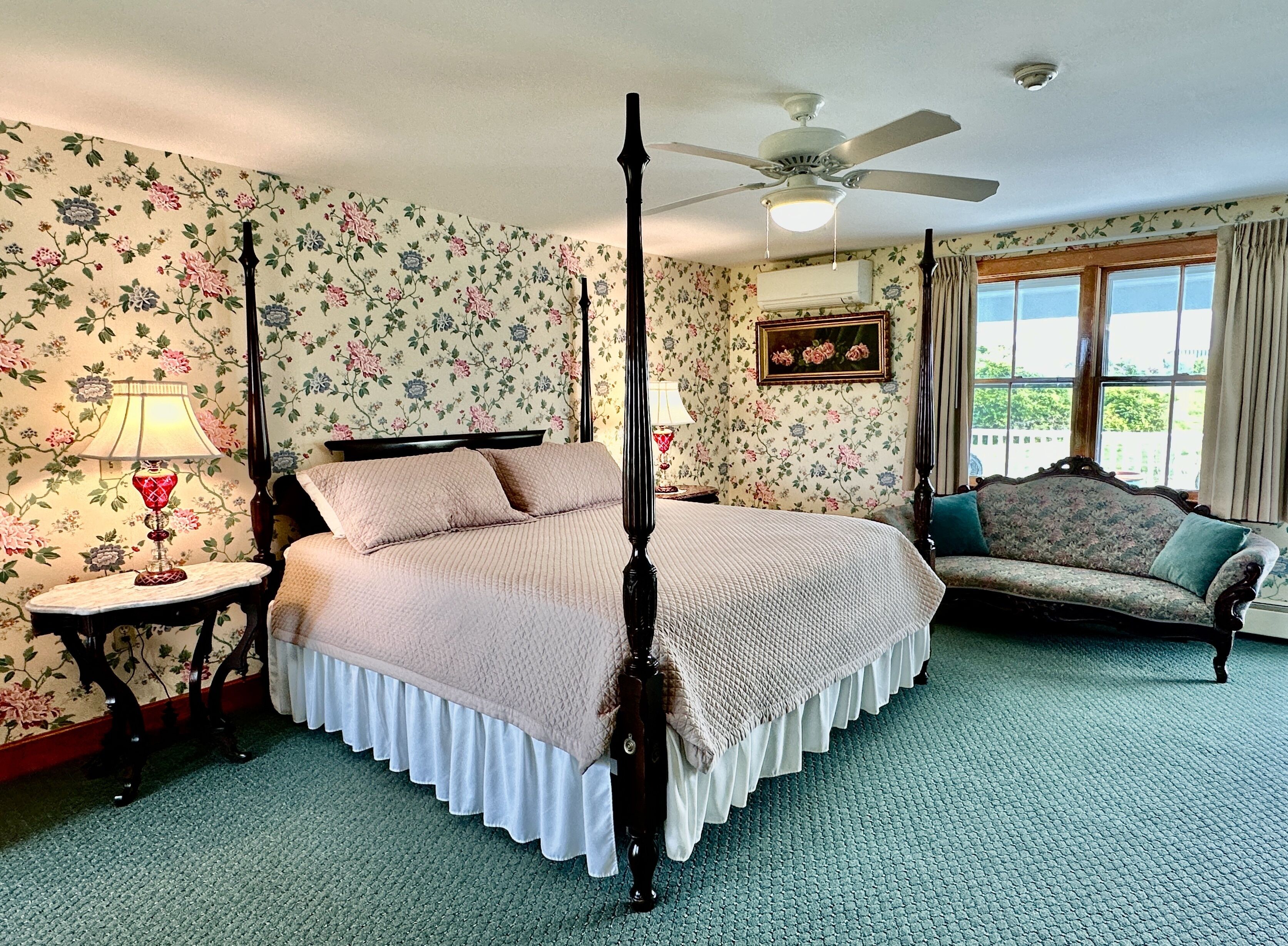 A four-poster bed with a white bedspread, a nightstand with a red lamp, a floral wallpaper, a ceiling fan, and a window with curtains.