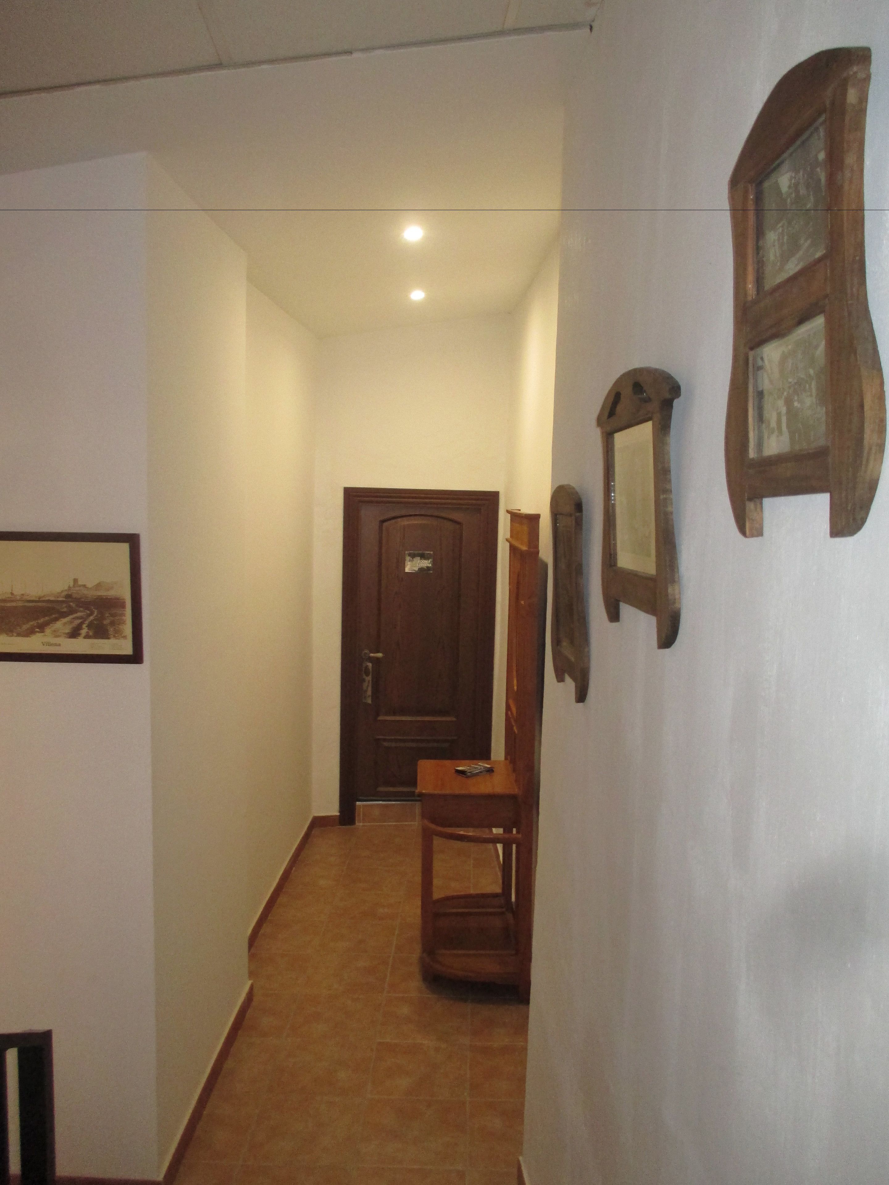 Un couloir étroit, avec une porte en bois au fond, une chaise et un tableau encadré au mur.