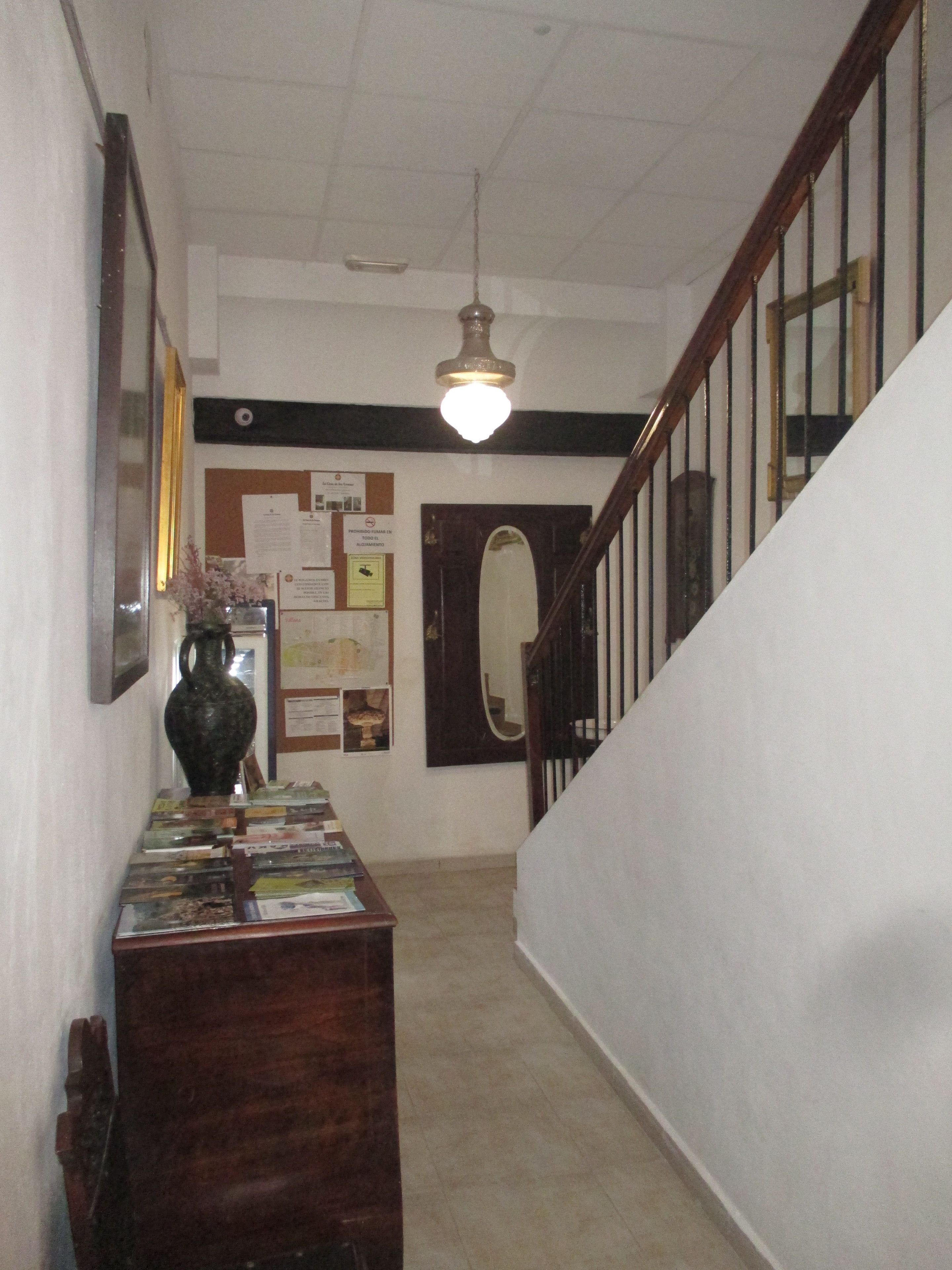 Un couloir avec un bureau en bois sur lequel sont disposés des livres et un vase, un panneau d’affichage où sont accrochées des annonces, et un escalier avec une rampe en bois.