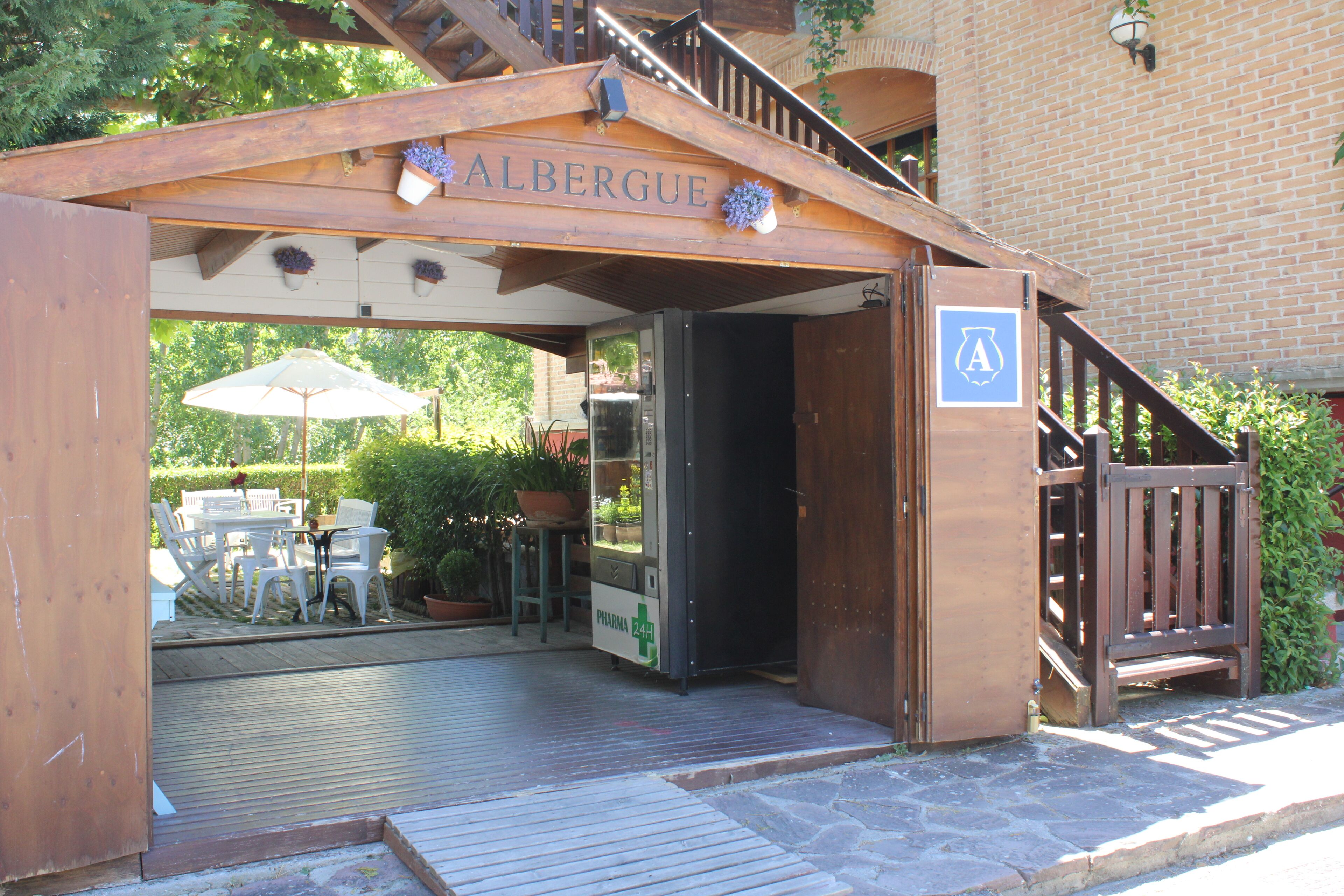 Entrada de madeira com uma placa que diz "ALBERQUE" e uma rampa acessível a cadeiras de rodas que leva a um pátio com área de estar ao ar livre e plantas em vasos.