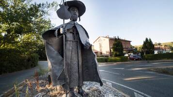 Eine Statue einer historischen Persönlichkeit mit Hut und Stab, auf einem felsigen Vorsprung neben einer Straße, im Hintergrund sind Gebäude zu sehen.