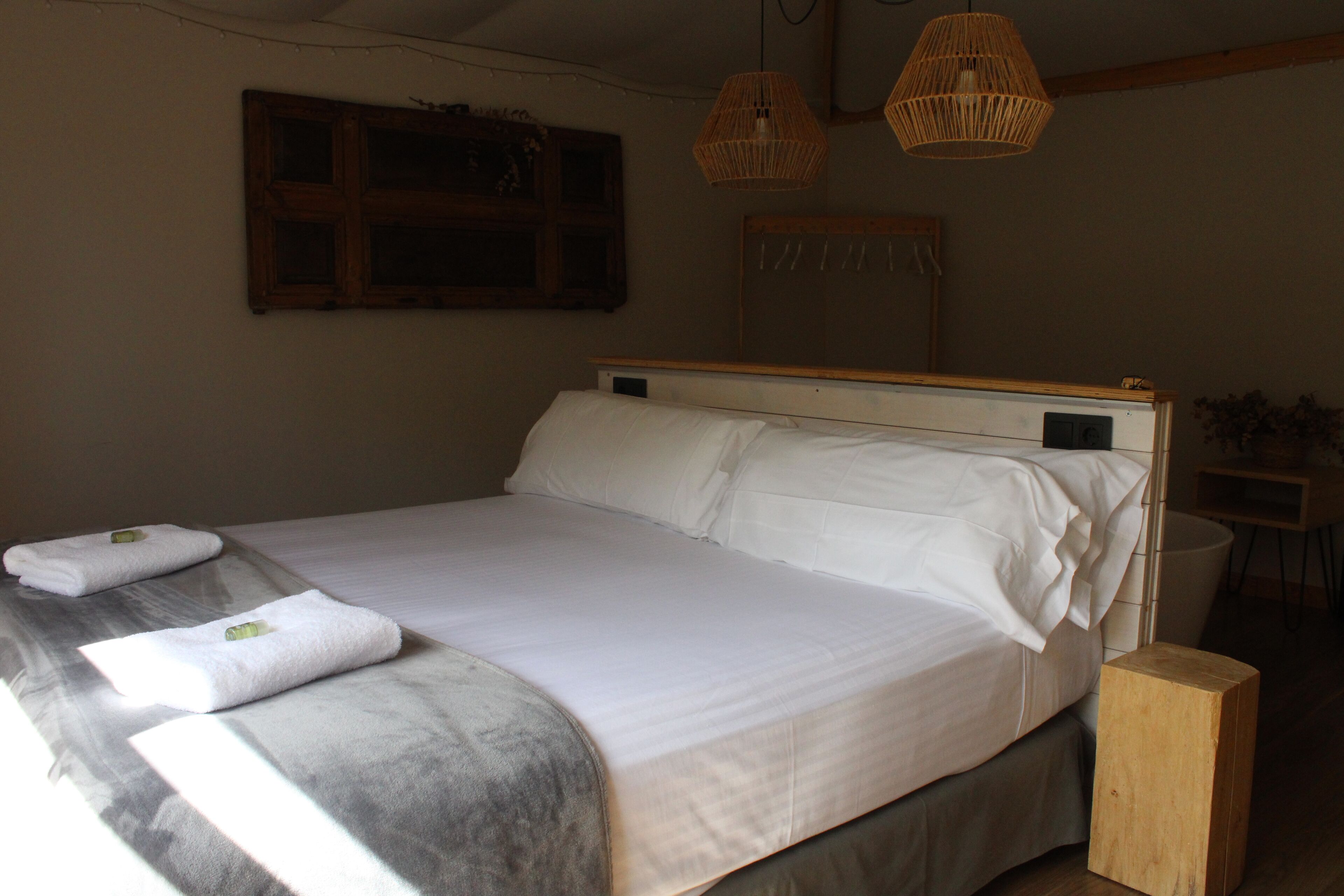 Una cama bien tendida con sábanas blancas y dos toallas, cabecera de madera y lámparas de mimbre.