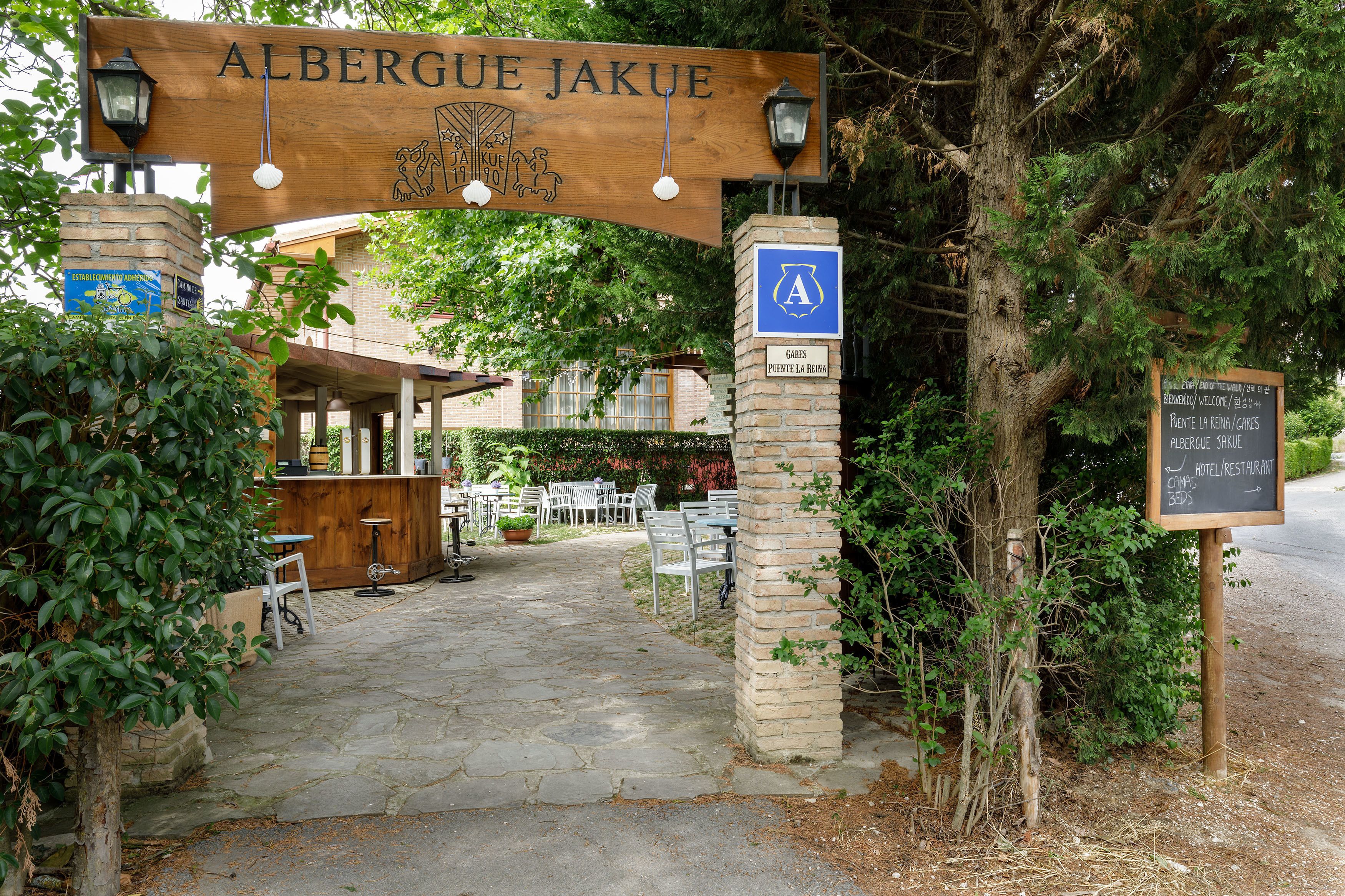 Un letrero de madera con 'ALBERGUE JAKUE' y un logotipo, un sendero de piedra que lleva a una zona de bar con mesas al aire libre, y una señal que indica un área de estacionamiento.