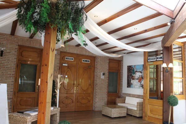 A lobby with wooden doors labeled "GLAMPING" and "ALBERQUE," wicker furniture, and a hanging plant. - Albergue Jakue - Hostel (Puente La Reina)