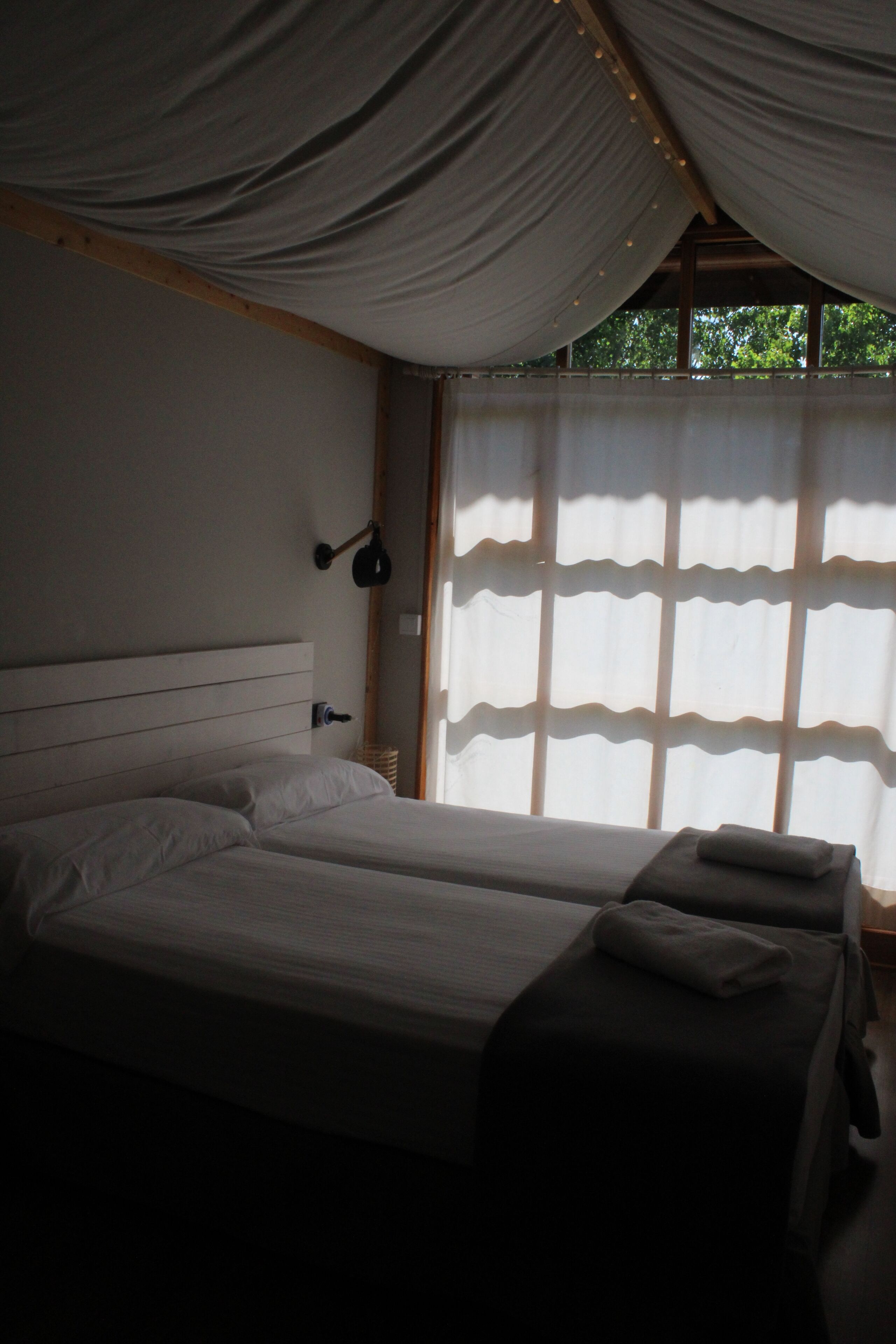 Habitación con dos camas, cabecero, lámpara de pared y ventana con cortinas traslúcidas.