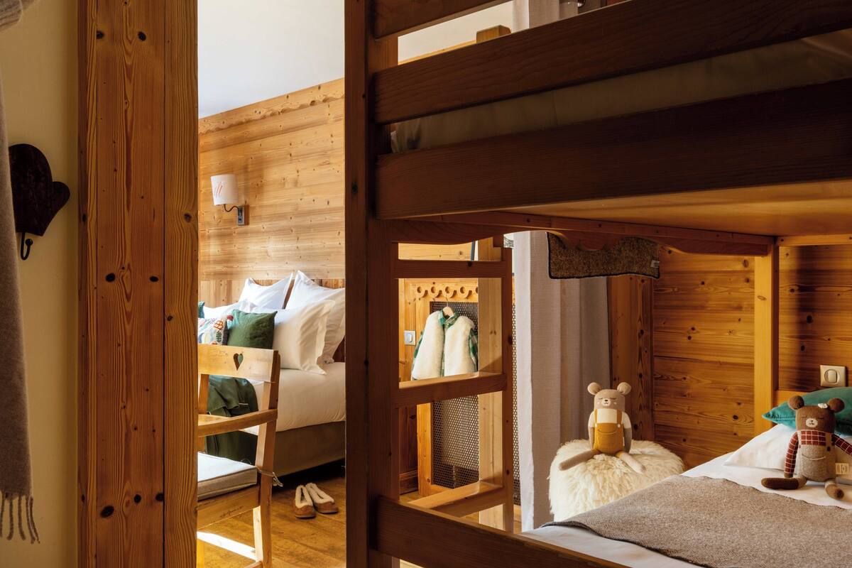 Une chambre avec un lit superposé en bois, une tête de lit en bois, une petite étagère avec quelques objets et un ours en peluche sur le lit.
