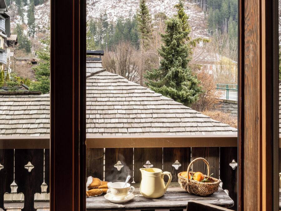 Uma varanda com uma mesa posta para chá, uma cadeira e vista para uma paisagem montanhosa coberta de neve.