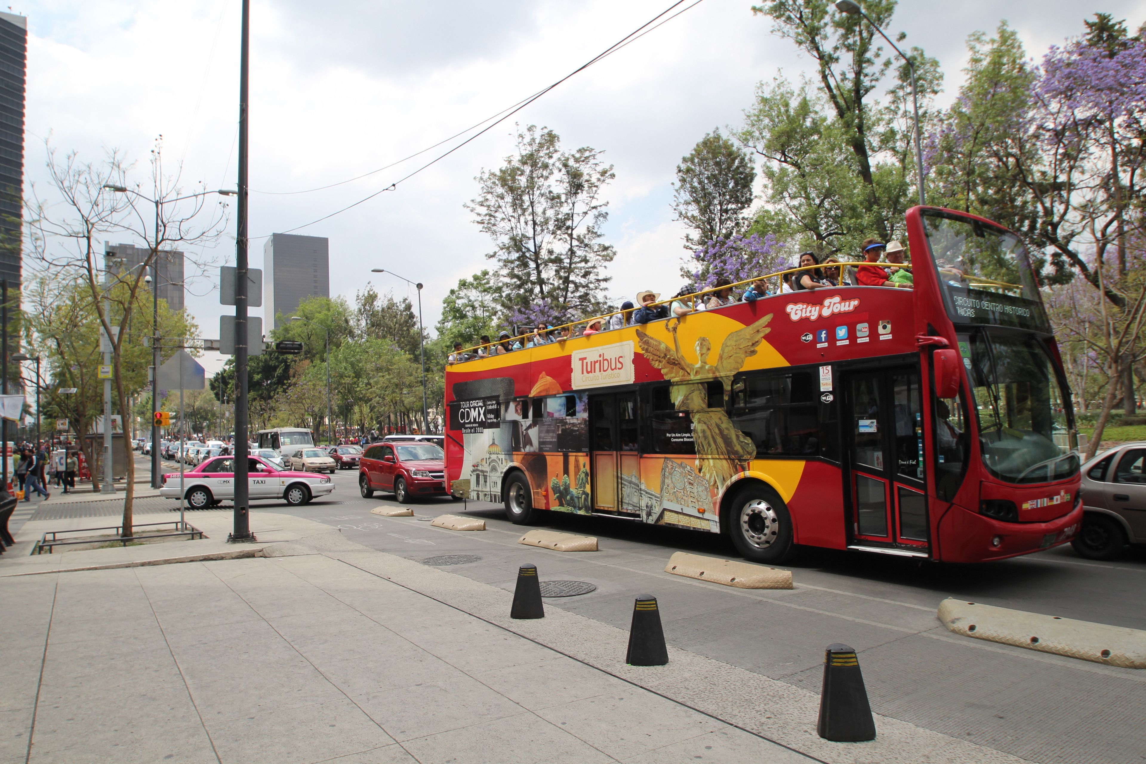 A red double-decker bus with "TURIBUS" branding and "City Tour" text, traveling on a city street with buildings and trees in the background.