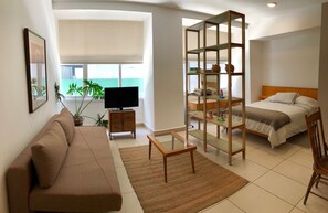 A modern hotel room with a sofa, a glass-top table, a bed with a wooden headboard, a TV, a bookshelf, and a view of the ocean. - Apartment Downtown 304 (Mexico City)