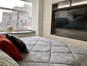 Un dormitorio moderno con una cama grande, un televisor montado en la pared y una ventana con vistas a edificios.