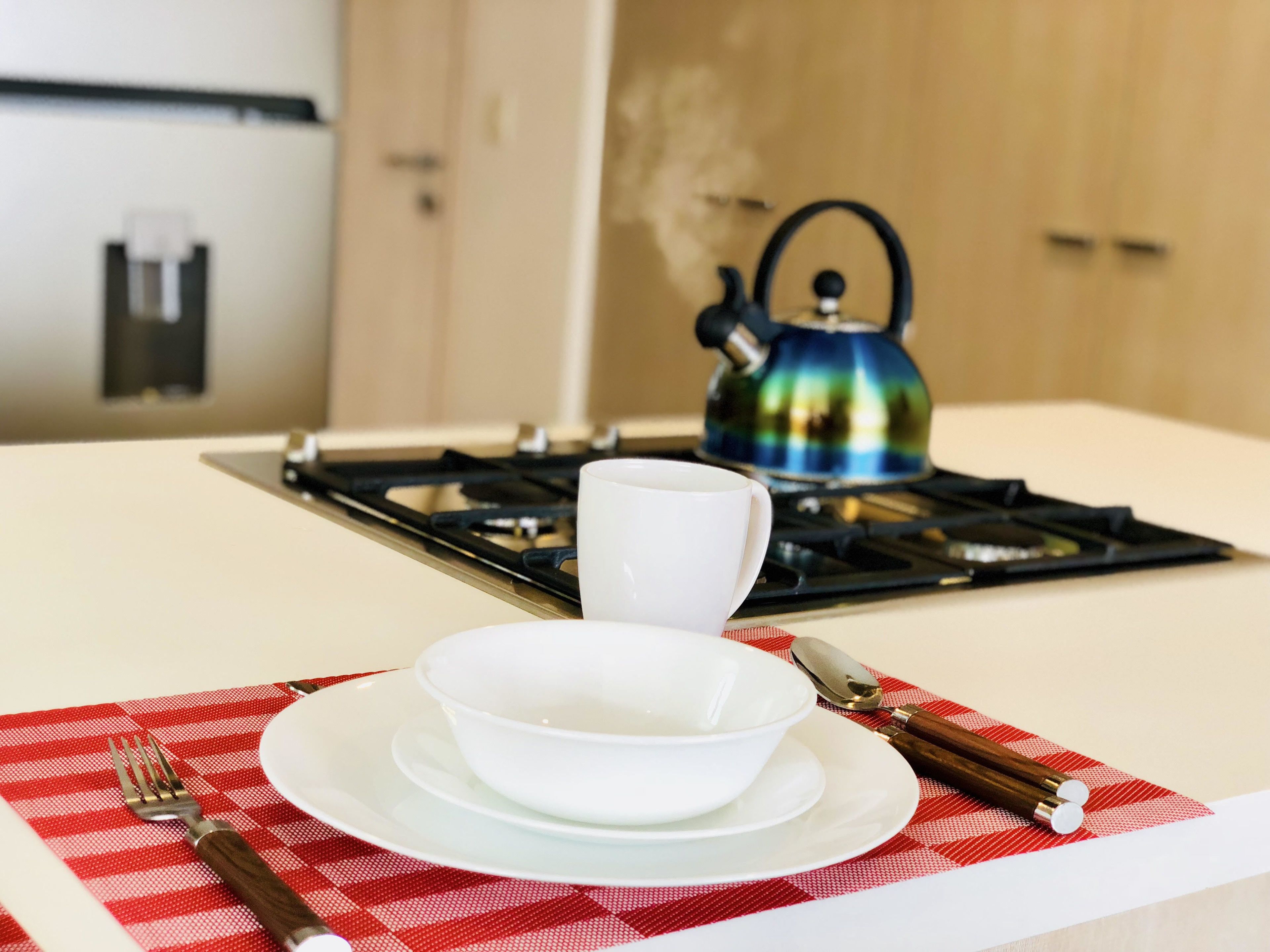 A kitchen with a gas stove, a kettle, a cup, a bowl, a fork, and a spoon on a red and white checkered placemat.