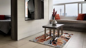 Una sala de estar moderna con un televisor de pantalla plana montado en la pared, una mesita con un florero y vistas a un paisaje urbano a través de amplios ventanales.