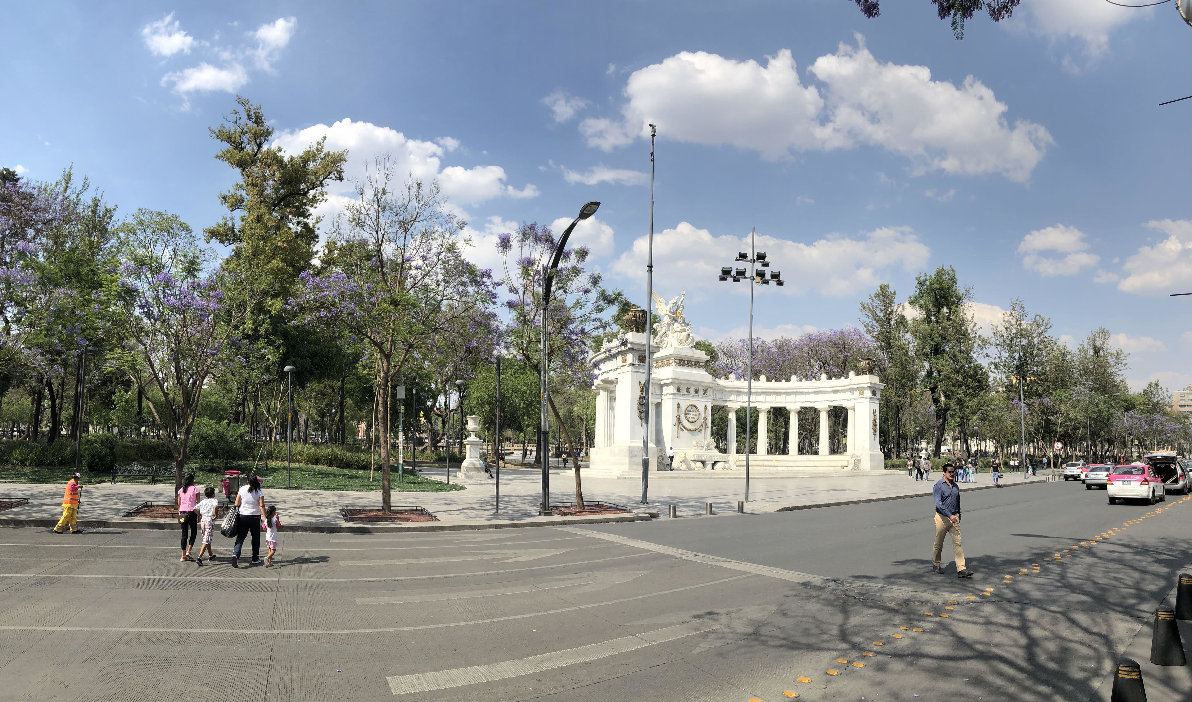 A public square with a central monument, surrounding trees, and a crosswalk.