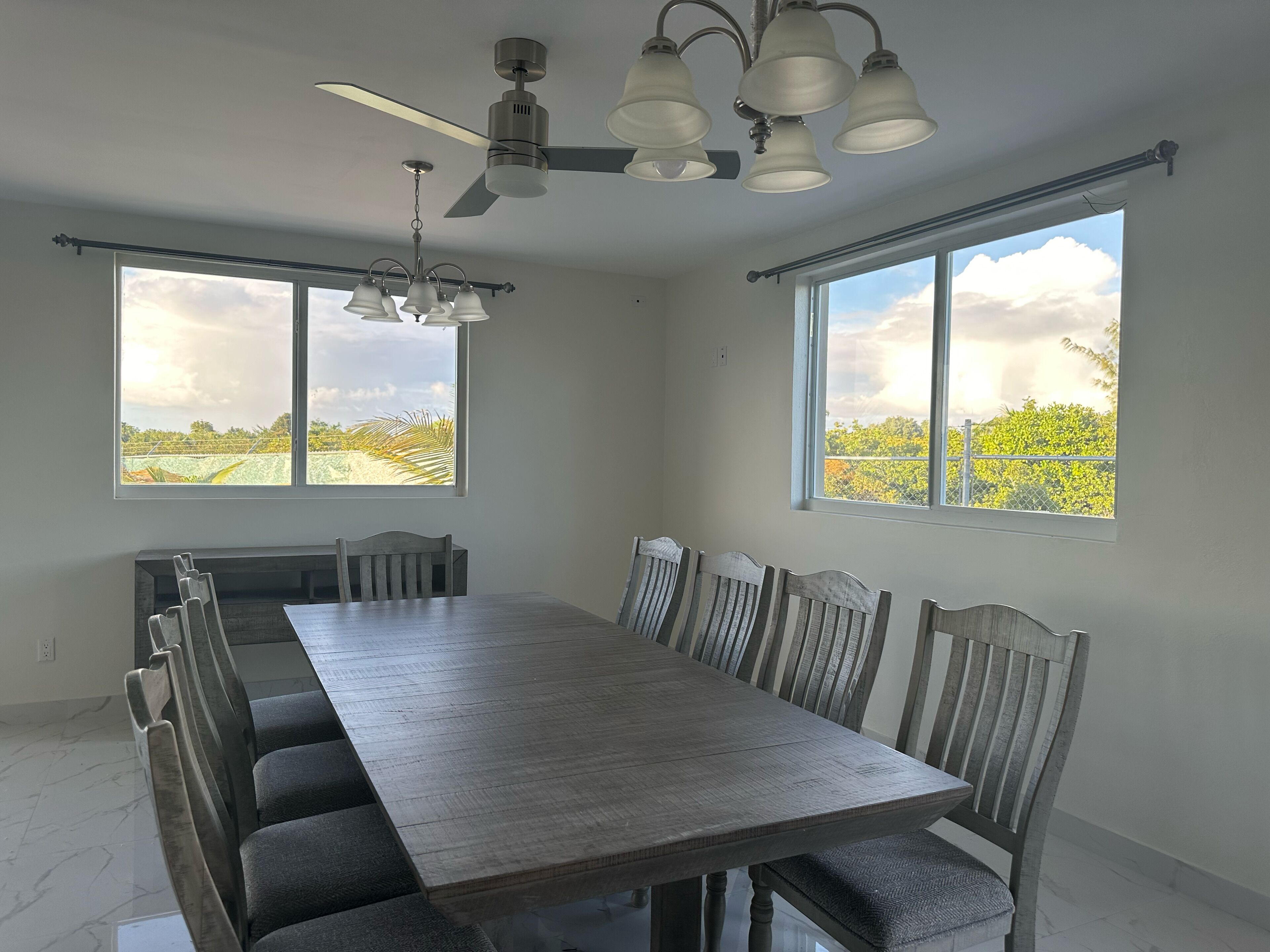 Un comedor con una mesa grande y sillas, un ventilador de techo y ventanas amplias con vistas a la vegetación.