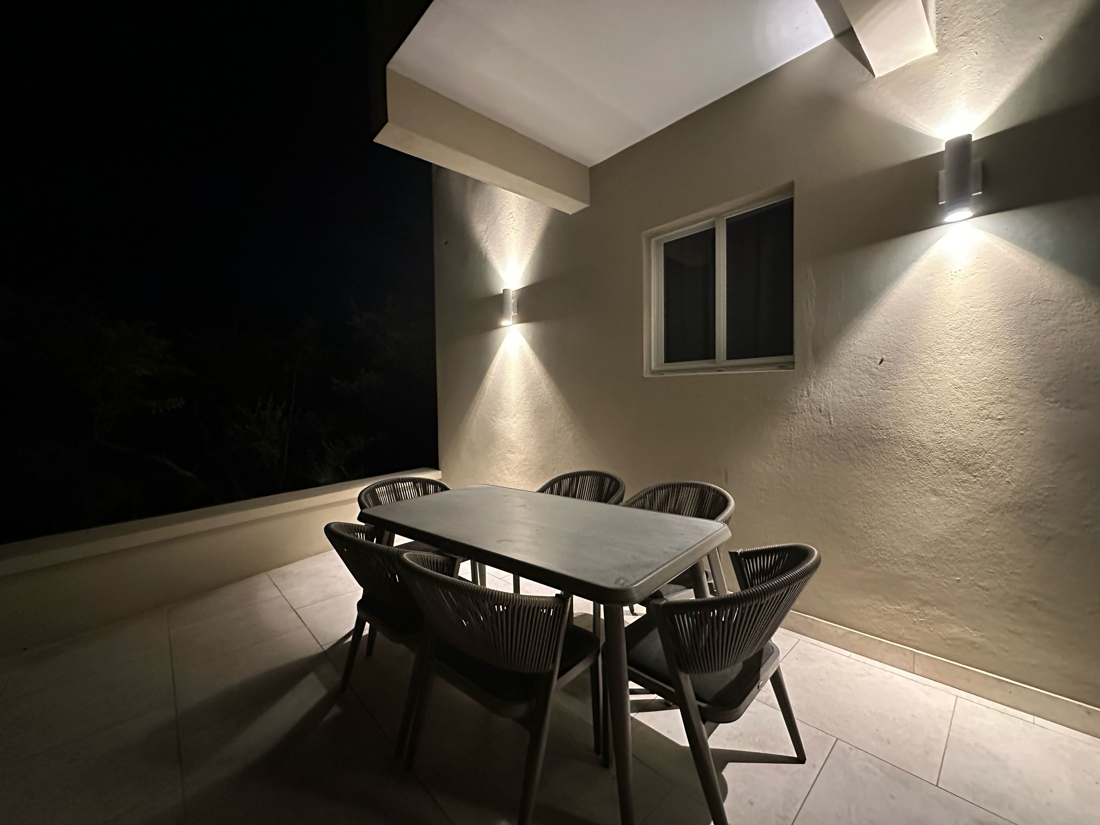 Une terrasse extérieure avec une table et des chaises, une fenêtre et des luminaires fixés au mur.