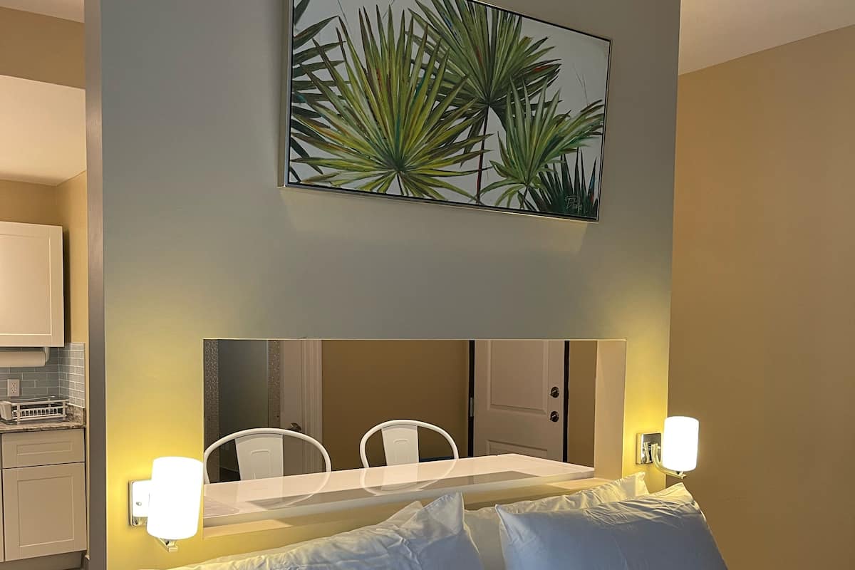 Een hotelkamer met een bed, nachtlampjes, een spiegel, een klein tafeltje en een keuken in de achtergrond.