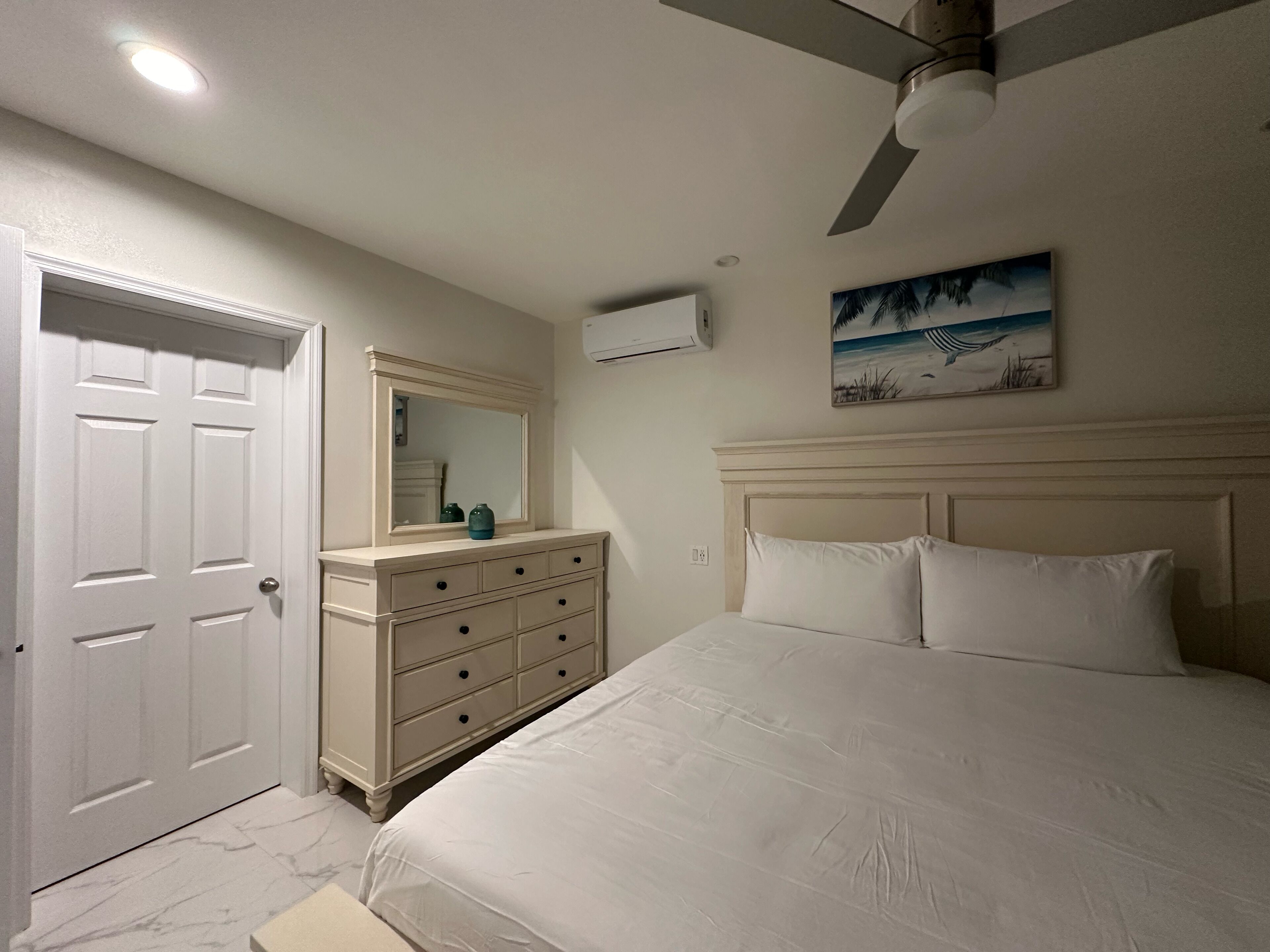 A bedroom with a bed, a dresser, a ceiling fan, and a painting on the wall.