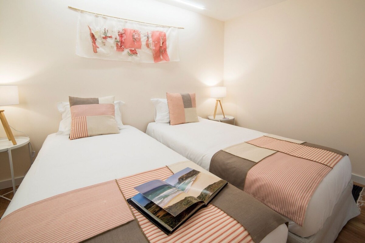 A hotel room with two beds, each with a pillow and a bedside table. A magazine is placed on the bed.