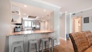 A modern kitchen with white cabinetry, a marble countertop, and stainless steel appliances. There are three bar stools and a wicker sofa in the living area. - Updated direct oceanfront 2/2 with a heated beachfront pool and beach access boardwalk (New Smyrna Beach)