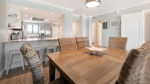 A dining area with a wooden table and wicker chairs, a kitchen in the background, and a ceiling light fixture. - Updated direct oceanfront 2/2 with a heated beachfront pool and beach access boardwalk (New Smyrna Beach)