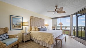 A bedroom with a large bed, a wicker chair, a bedside table, a ceiling fan, and a view of the ocean.