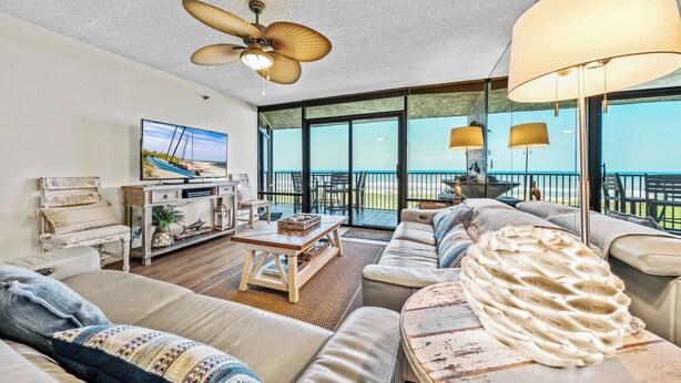 A spacious living room with a large sofa, a coffee table, and a television. There is a balcony with a view of the ocean.