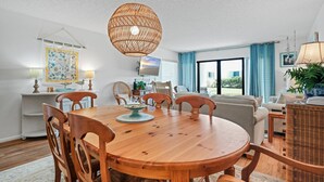 A dining area with a wooden table and chairs, a hanging light fixture, and a view of the outdoors through large windows. - Ground floor 2/2 condo with a large patio, just steps from the pool and beach access (New Smyrna Beach)