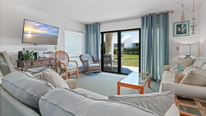 A modern living room with a sofa, armchairs, a coffee table, and a TV. There is a sliding glass door leading to a balcony. - Ground floor 2/2 condo with a large patio, just steps from the pool and beach access (New Smyrna Beach)
