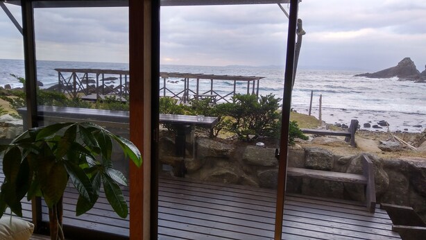 Una terraza de madera con cerramiento de vidrio que da a una playa rocosa y al mar.