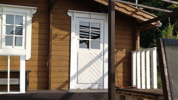 A wooden cabin with a white door and windows, a wooden deck, and a pergola structure.