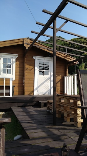 A wooden cabin with a white door and windows, a wooden deck, and a pergola structure.