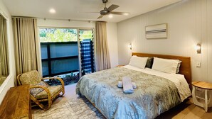 A bedroom with a large bed, a ceiling fan, a chair, a small table, and a view of greenery outside. - Banksia Luxury Villa (Margaret River)