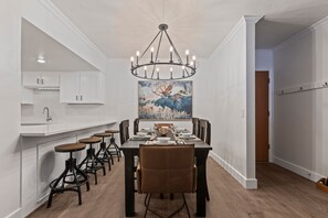 A dining area with a long table set for six, surrounded by six chairs. A chandelier hangs above the table, and a painting of a moose is on the wall behind the table.
