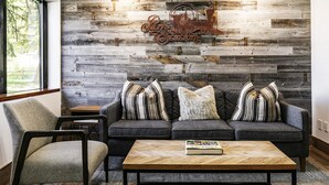 Een moderne woonkamer met een grijze modulaire bank, gestreepte kussens, een houten salontafel en een rustieke wand met een 'Park & Rest' bordje.