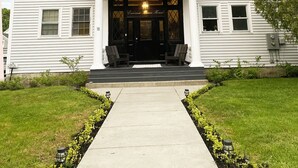 A two-story white building with a central entrance, two side entrances, and a front porch with chairs. - 3500 SF Original School transformed into an artful,  gorgeous village home, yard (Bar Harbor)
