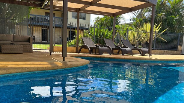 Une piscine avec des chaises longues, un espace de détente couvert et un bâtiment en arrière-plan.