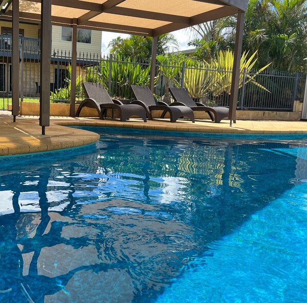 A swimming pool with lounge chairs, a covered seating area, and a building in the background.