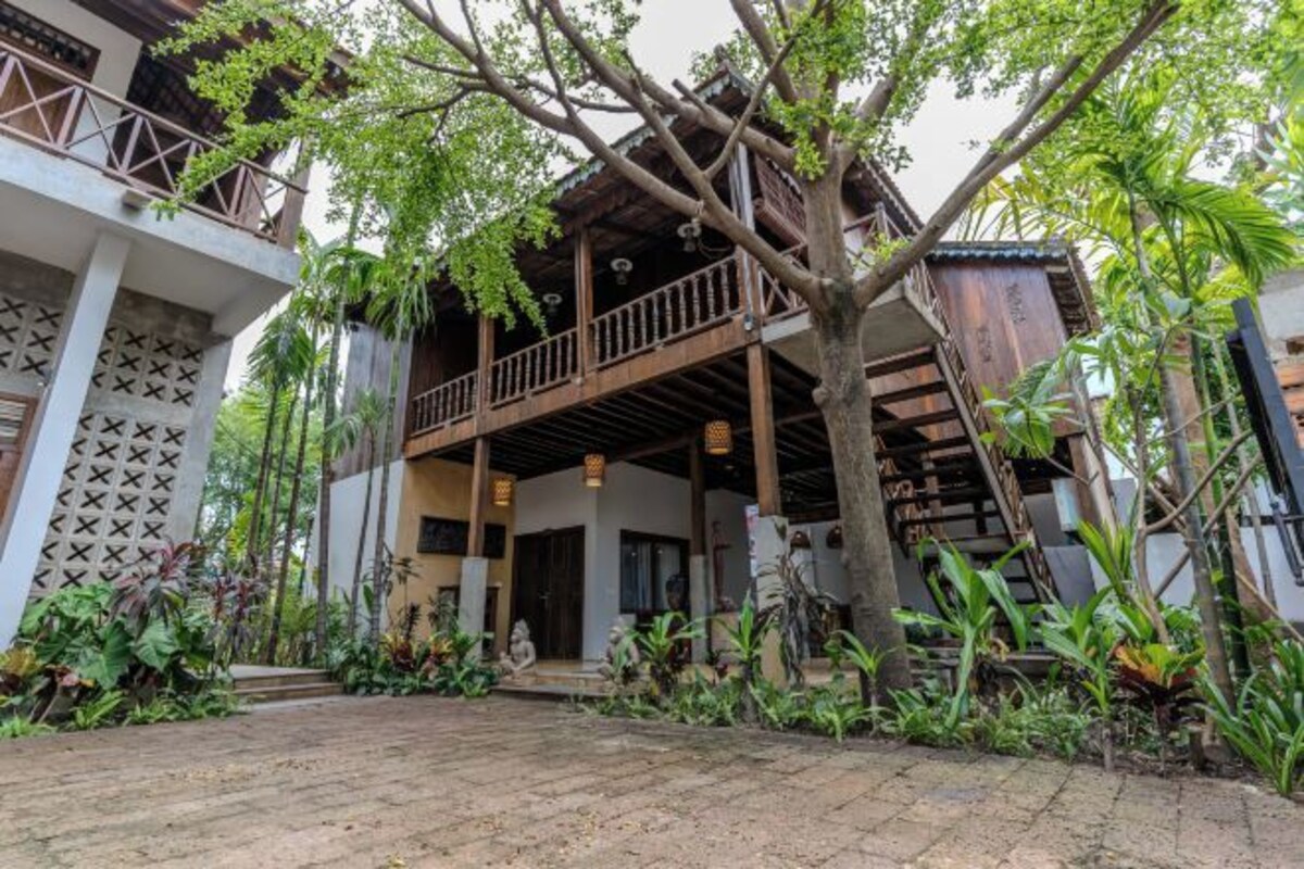 A traditional wooden building with a balcony, surrounded by lush greenery and a paved walkway.