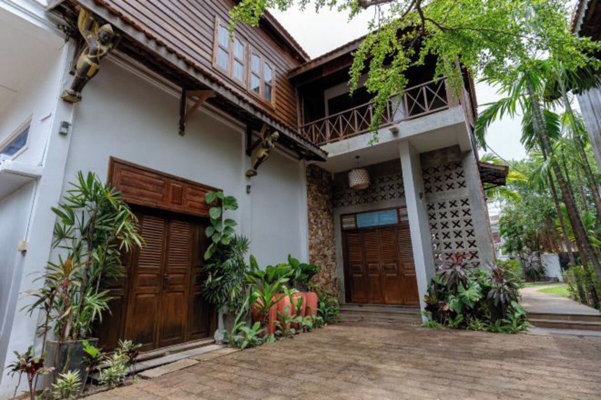 A traditional house with a wooden balcony, large wooden doors, and lush greenery around the entrance.