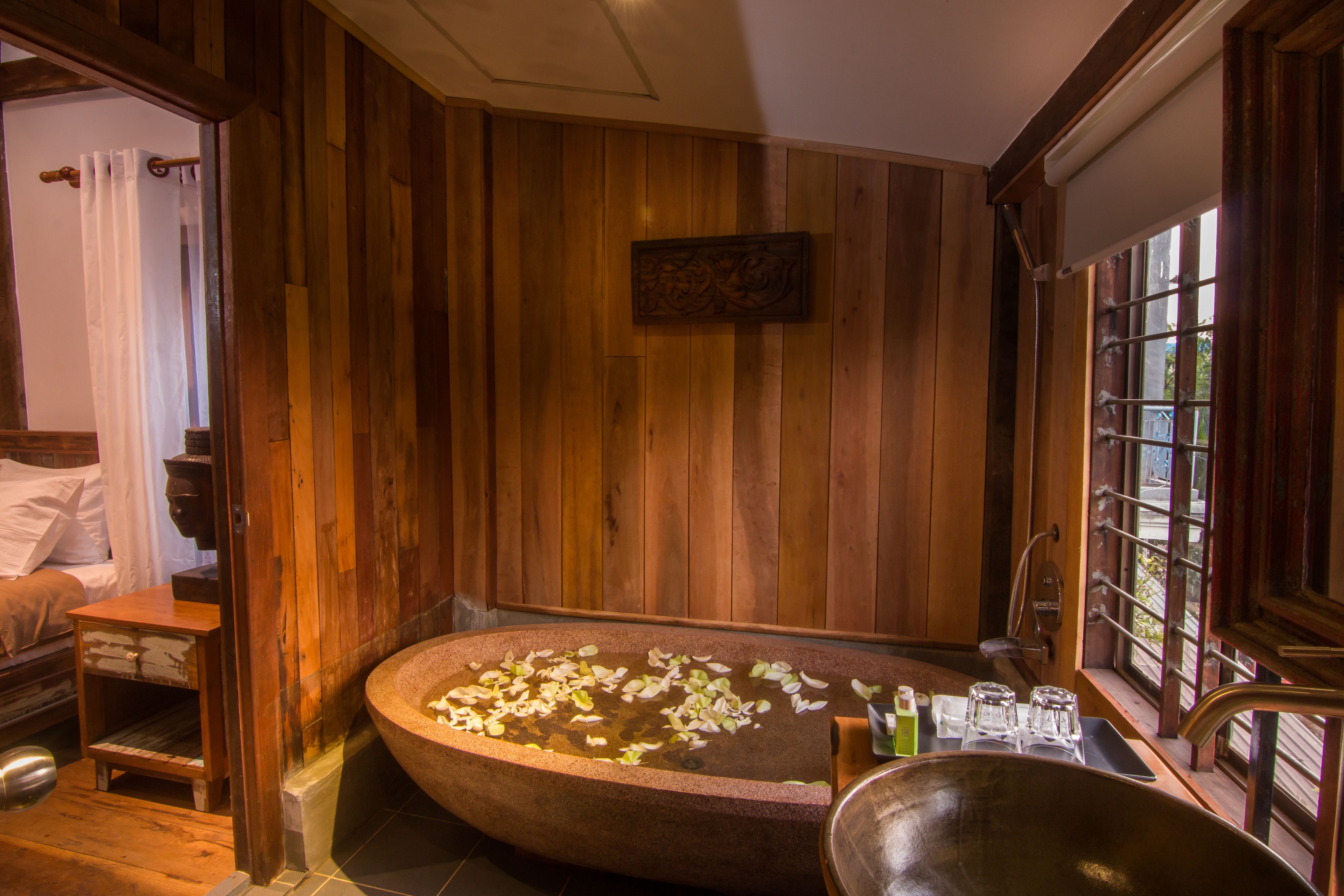 A wooden room with a large bathtub filled with water and flower petals, a wooden bench, and a window with a view.