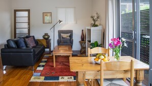 A living room with a black sofa, a wooden coffee table, a dining table with chairs, a red rug, and a window with blinds. - St Clair Cottage Peaceful romantic country getaway  (Margaret River)