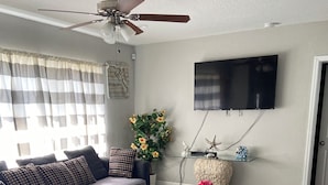 Un salon comprenant un canapé d’angle, une table basse en verre, un ventilateur de plafond et un téléviseur à écran plat fixé au mur.
