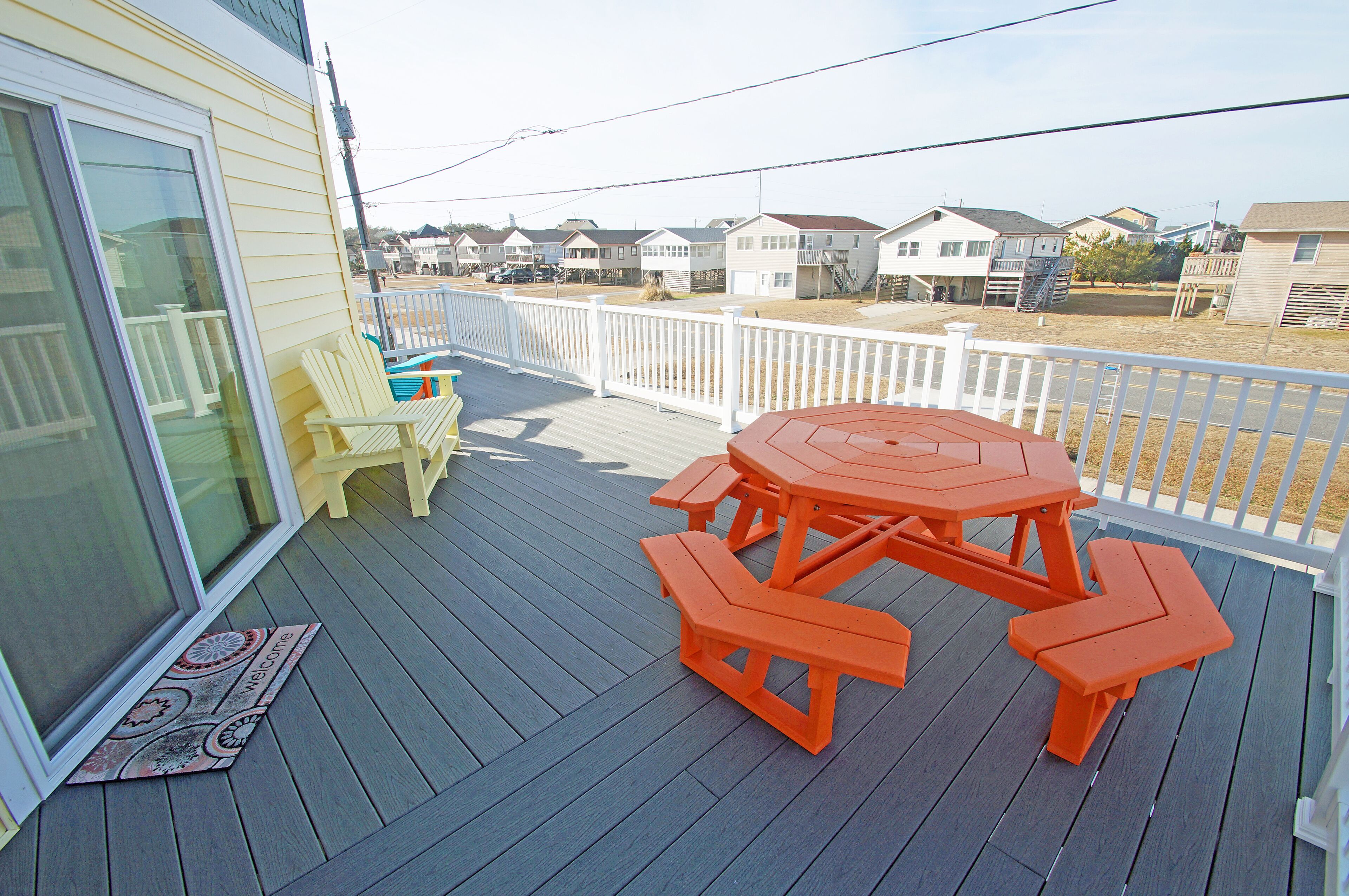 A wooden deck with a picnic table and chairs, a welcome mat, and a view of houses in the background.