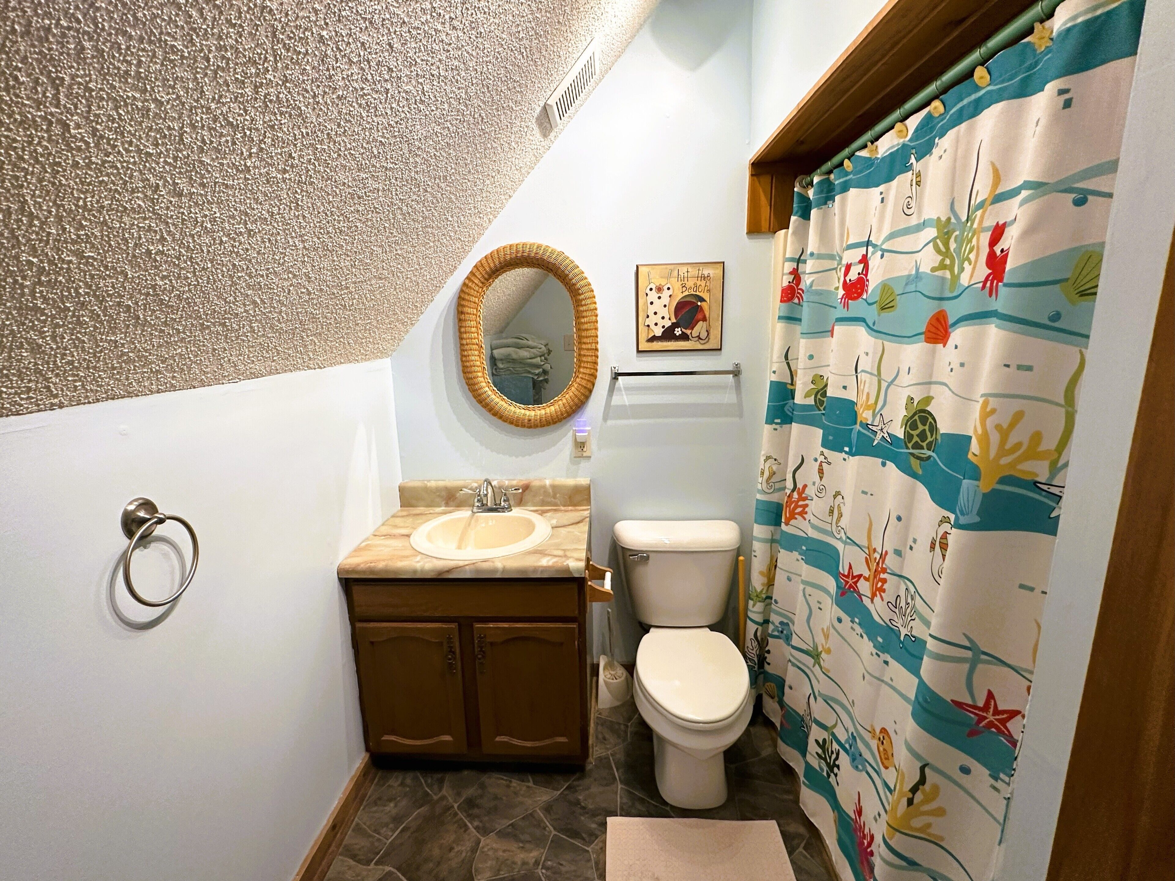 Une salle de bain avec un lavabo en marbre, des toilettes, un miroir en osier et un rideau de douche sur le thème de la mer.