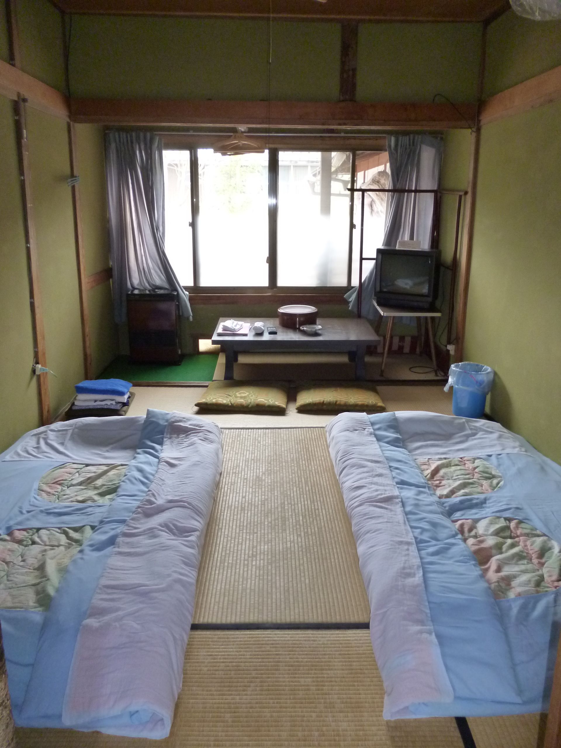 A traditional Japanese-style room with tatami mats, two tatami beds, a futon, a television on a stand, and a small table with a kettle and cups.