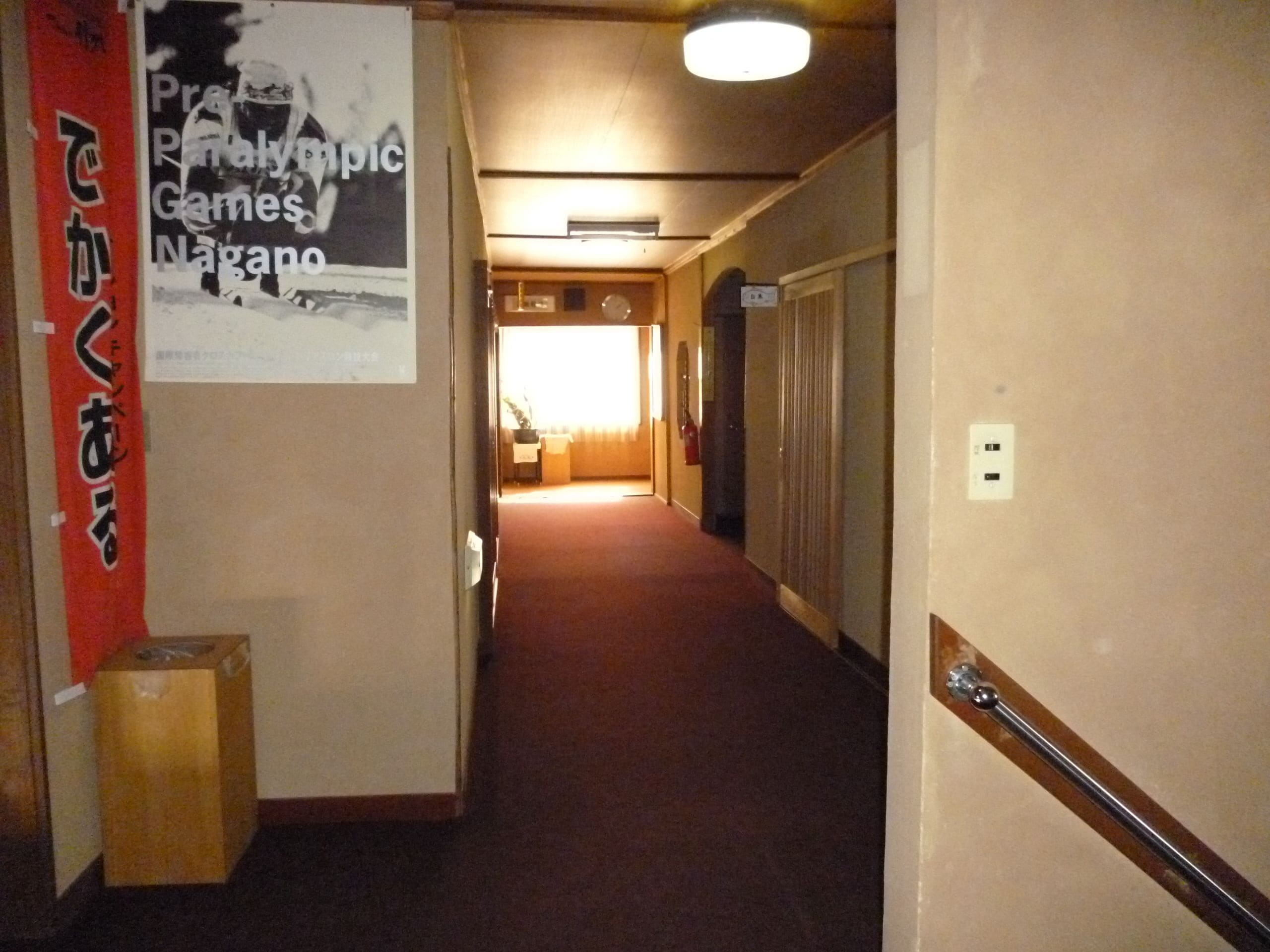 Um corredor de hotel com um cartaz na parede, uma caixa de madeira e uma porta no final.