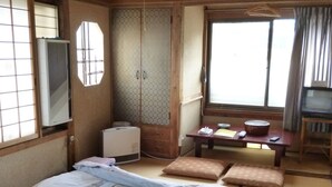 Ein traditionelles japanisches Schlafzimmer mit Tatami-Boden, einem großen Bett mit gemusterten Bettwaren, einem Kleiderschrank mit Schiebetüren, einem Fernseher und einem kleinen Tisch mit Kissen.