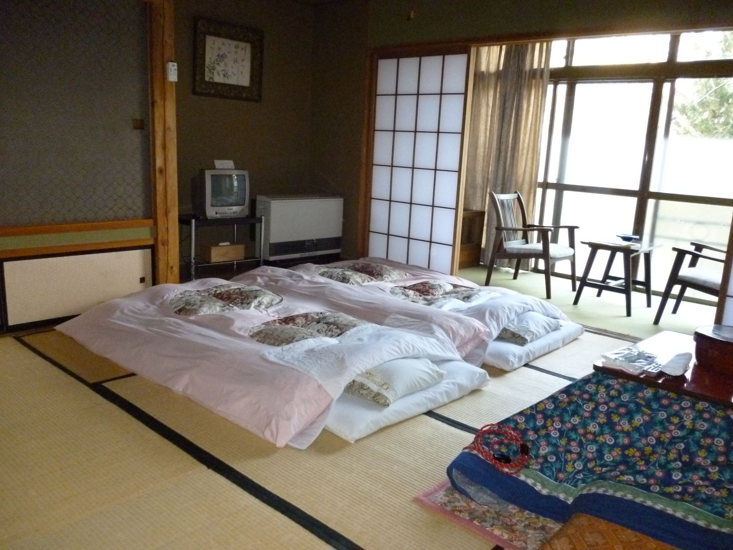 Een traditionele Japanse kamer met tatami vloerbedekking, een futonbed, een televisie en een schuifdeur naar een balkon met stoelen.
