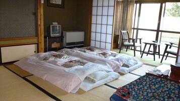 Een traditionele Japanse kamer met tatami vloerbedekking, een futonbed, een televisie en een schuifdeur naar een balkon met stoelen.